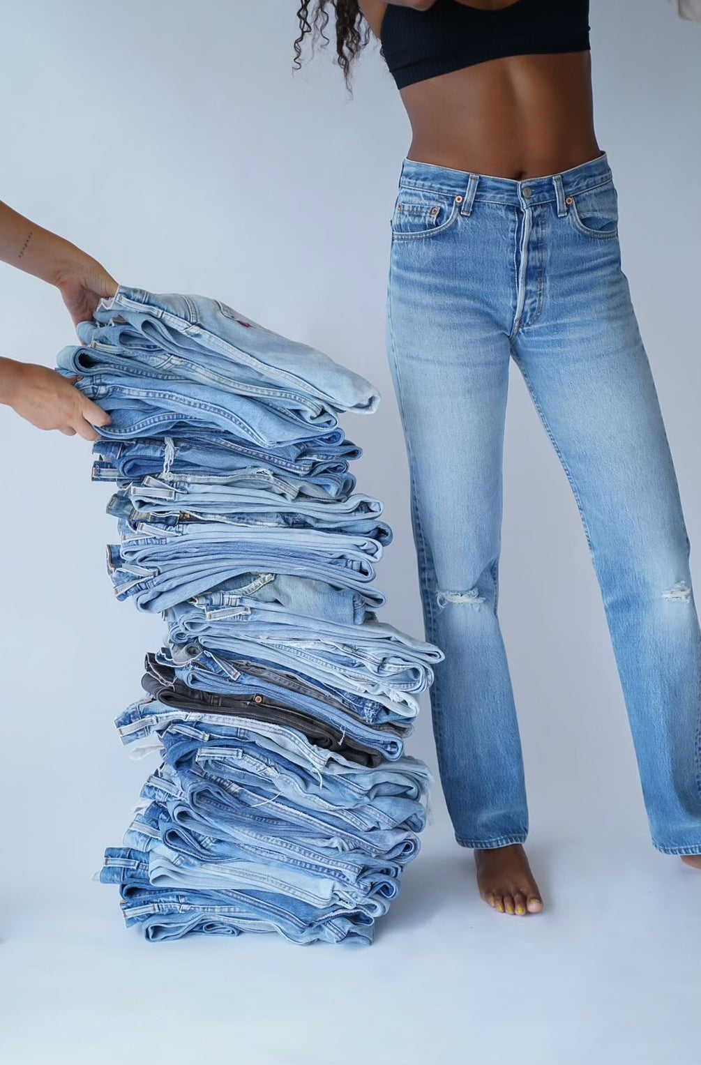 A model is pictured from the torso, down standing next to a stack of folded jeans. Two hands are in the frame stacking more pairs.